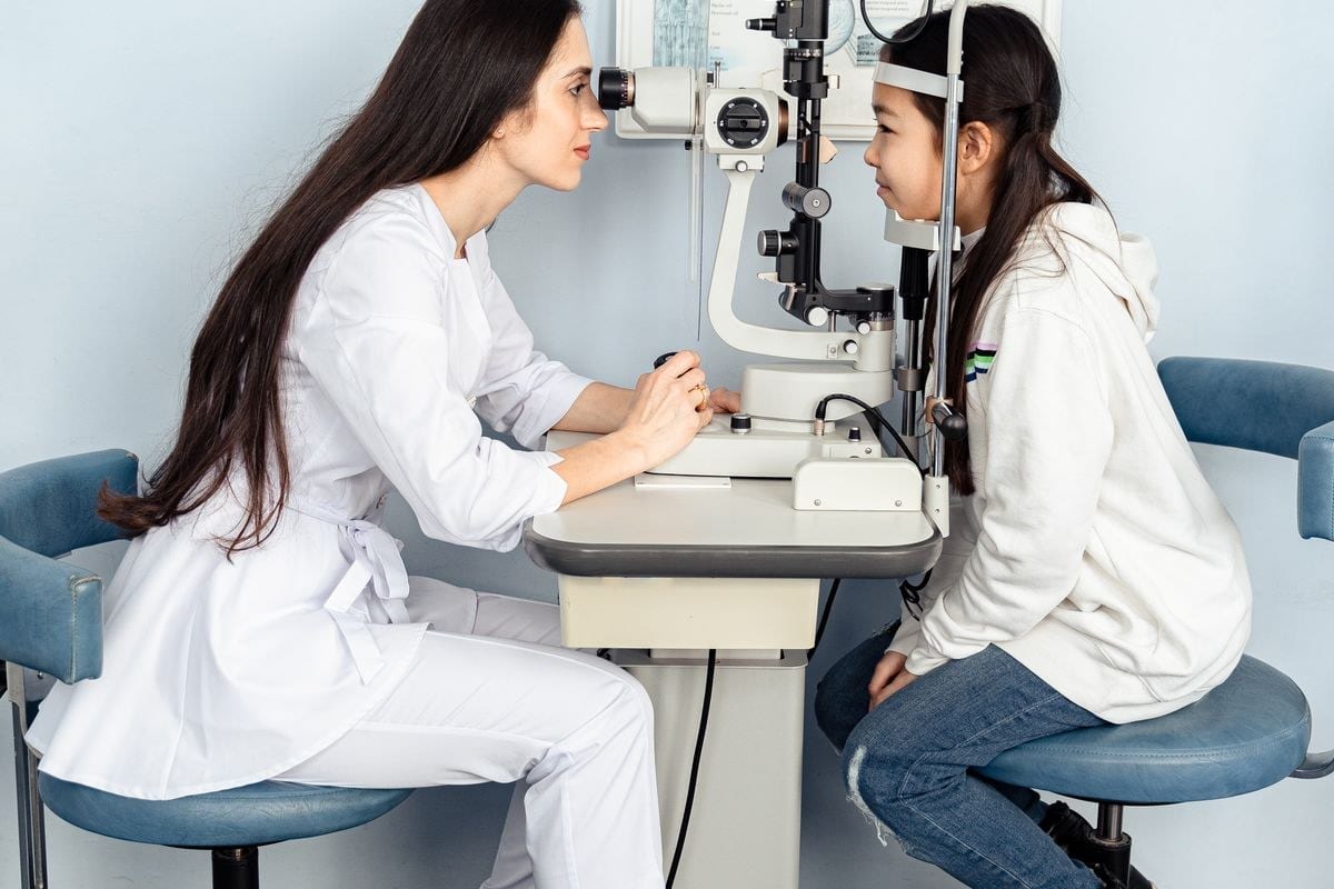 Beyaz önlüklü göz doktoru, biyomikroskop başında oturan çocuk hastanın gözlerini inceliyor
